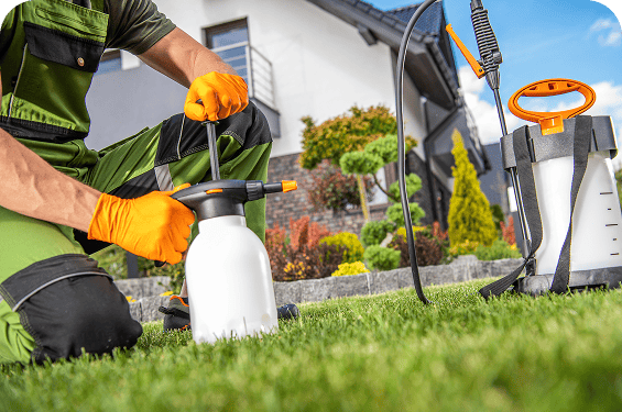 Person in green workwear and orange gloves prepares a garden sprayer on a lawn, demonstrating coastal termite and pest control techniques, with landscaping and a house visible in the background.